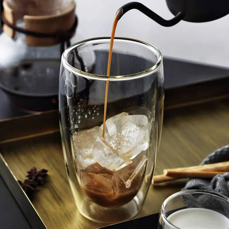 Iced coffee being poured into a glass with ice cubes on a wooden tray.