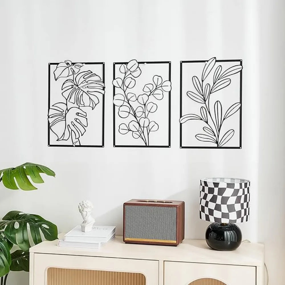 Three black and white leaf-patterned wall art pieces on a white wall above a white cabinet with decorative items.