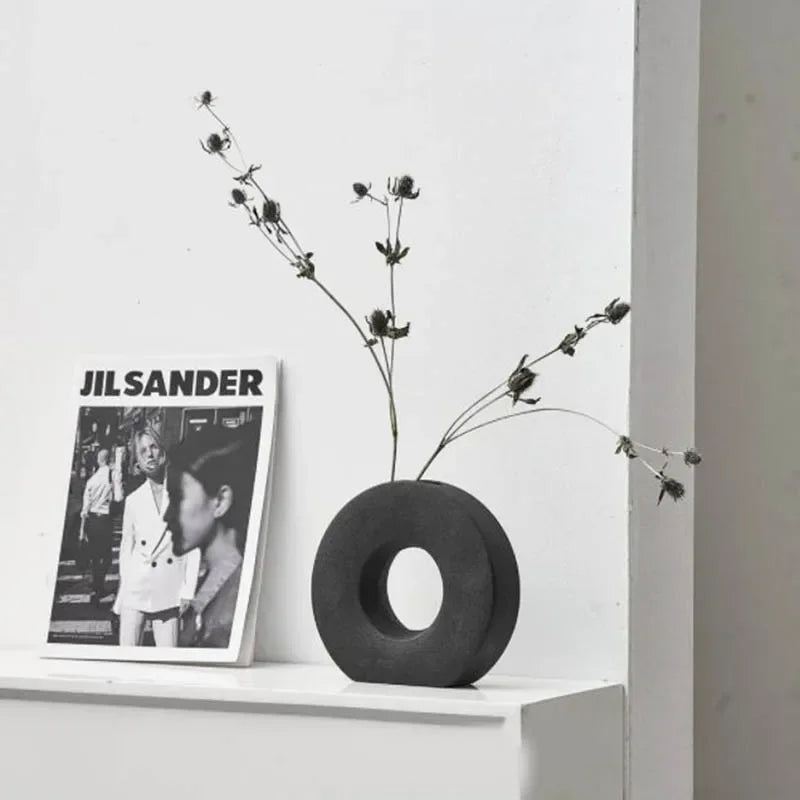 Black vase with dried branches on a white shelf next to a Jil Sander poster.