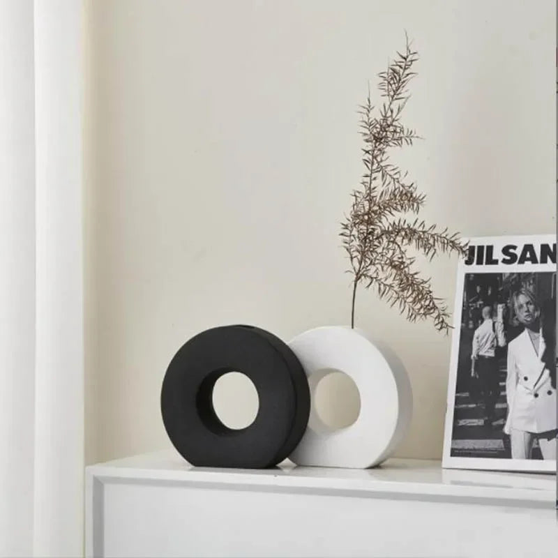Two decorative vases on a white surface with a plant and a framed picture in the background.