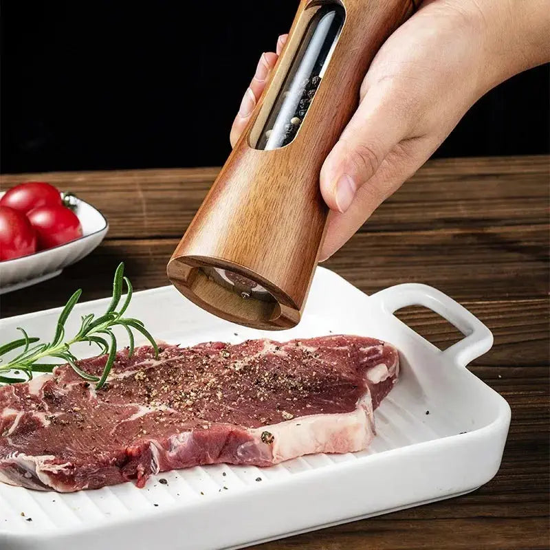 Hand using a wooden pepper grinder on a raw steak on a white plate with red peppers and rosemary in the background.