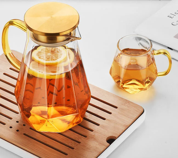 Glass teapot with gold lid and lemon slices, accompanied by a glass mug with tea on a wooden coaster.