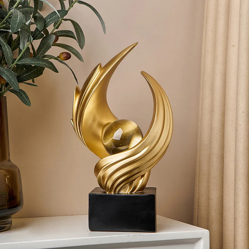 Gold abstract sculpture on a black base with a plant and curtain in the background